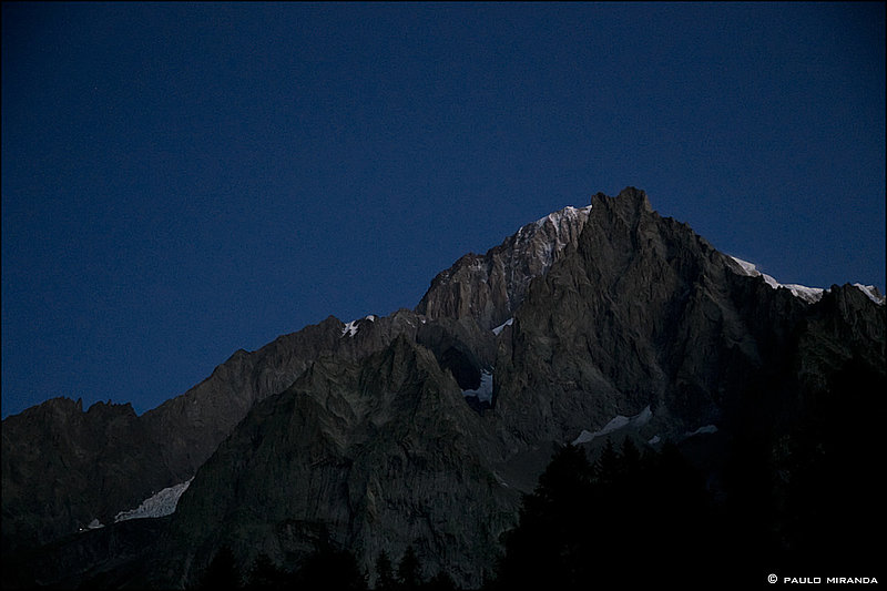 Em primeiro plano, a Aiguille Noire de Peuterey (3.773 m) visto no início da manhã do Col Chècrouit, e ao fundo, as encostas do Monte Branco. No canto inferior esquerdo, as luzes do Refúgio Monzino.